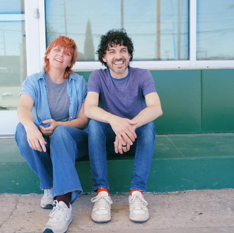 Zach and Amanda sit on the dark green step of a dark green building with large white-framed windows and a glass door. They're both wearing jeans, blue shirts, and white shoes, and are squinting at the sun. They look happy but also, perhaps, wonder what's going on.